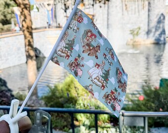 May include: A small flag with a white pole featuring a light blue background and a repeating pattern of cartoon characters, Christmas trees, and presents. The flag is held up in front of a castle and a body of water.