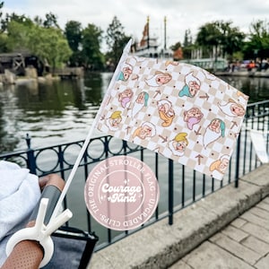 May include: A stroller flag featuring a pattern of cartoon dwarves on a tan and white checkered background. The flag is held by a white plastic clip. The text on the flag reads "The Original Stroller Flag, Courage + Kind, Upgraded Clips."