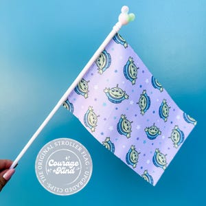 May include: A small stroller flag featuring a repeating pattern of green, three-eyed aliens on a lavender background with blue stars. The flag is attached to a white pole with a Mickey Mouse-shaped topper. The flag reads "Courage + Kind".