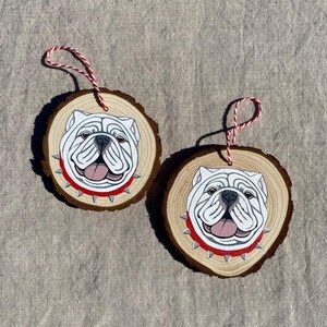 May include: Two wooden ornaments with painted images of white bulldogs wearing red collars with silver studs. The ornaments are round and have a rustic wood grain texture.