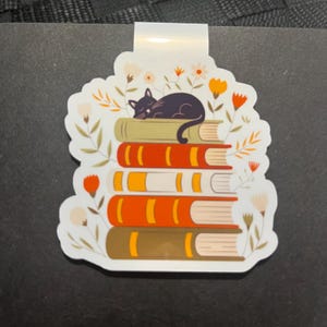 Puede incluir: Marcapáginas blanco con un diseño de un gato negro durmiendo sobre una pila de libros. Los libros son en tonos naranja, verde y marrón, con detalles florales en naranja y blanco. El marcapáginas tiene una pestaña blanca.