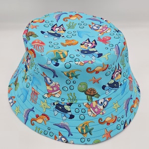 May include: A light blue bucket hat with a repeating pattern of cartoon sea creatures and characters. The design includes fish, turtles, jellyfish, and a cartoon dog character. The hat is designed for children.
