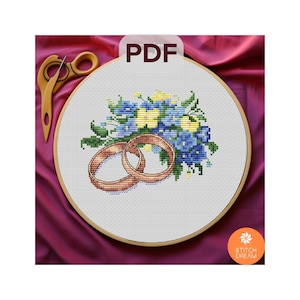 Puede incluir: Un patrón de punto de cruz con dos anillos de boda dorados y un ramo de flores azules y amarillas. El patrón se muestra en un aro de bordado blanco.