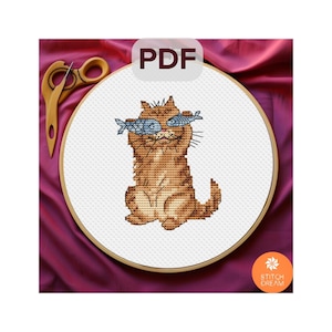 Puede incluir: Patrón de punto de cruz de un gato atigrado marrón sosteniendo dos peces azules frente a sus ojos. El diseño está en un bastidor de bordado de madera. La palabra "PDF" está en la esquina superior derecha. Unas tijeras de madera están arriba a la izquierda.