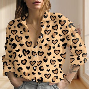 May include: A button-down shirt with a tan background and a pattern of black and brown hearts and leopard spots. The long-sleeved shirt has a collared neckline and is unbuttoned at the top.
