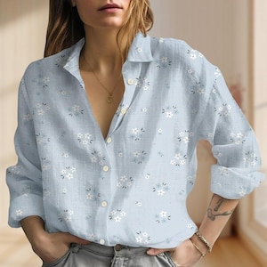 May include: A light blue button-down shirt with a floral pattern. The shirt features white and light blue flowers scattered across the fabric. The long-sleeved shirt has a classic collar and buttoned cuffs.