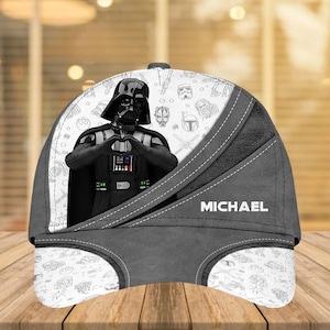 May include: A baseball cap featuring Darth Vader making a heart shape with his hands. The cap is primarily gray and white, with the name "MICHAEL" printed on the side. The design includes Star Wars character illustrations.
