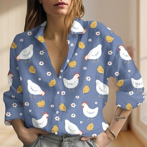 Puede incluir: Camisa azul claro con botones con un estampado repetido de gallinas blancas, pollitos amarillos y pequeñas flores blancas. La camisa de manga larga tiene un cuello clásico y está hecha de una tela suave y ligera.