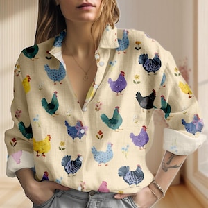 May include: A button-down shirt with a cream-colored background and a repeating pattern of colorful chickens and small flowers. The chickens are in various colors, including blue, purple, yellow, and green. The shirt has long sleeves with rolled-up cuffs.