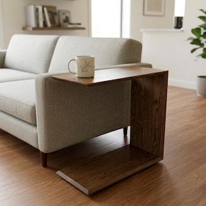 May include: A C-shaped side table crafted from dark brown wood with a visible grain pattern, positioned next to a light gray sofa. A white ceramic mug featuring a black line drawing of an olive branch sits on the flat top surface of the table.