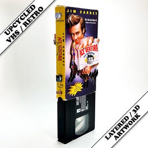 May include: Upcycled VHS tape artwork featuring the movie "Ace Ventura: Pet Detective" starring Jim Carrey. The artwork is layered and 3D, with the VHS tape case and movie cover visible. The text "Upcycled VHS / Retro" and "Layered / 3D Artwork" are also present.