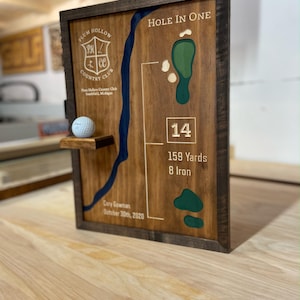 May include: Wooden golf-themed plaque with a dark frame. The plaque features a golf course layout, the words "Hole in One", and the text "Plum Hollow Country Club". A golf ball rests on a small shelf. The plaque includes the text "14", "159 Yards", and "8 Iron".