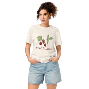 Op de afbeelding: Crèmekleurig T-shirt met een aquarel illustratie van wilde frambozen op een tak met groene bladeren. De tekst "Wild Raspberry" is eronder gedrukt in een bordeauxrode script. Het model draagt een blauwe spijkerbroek.