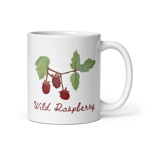 Op de afbeelding: Witte keramische mok met een aquarel illustratie van wilde frambozen. Het ontwerp toont drie rode frambozen, groene bladeren en de tekst "Wild Raspberry" in een sierlijk lettertype. Een klassieke koffiemok.