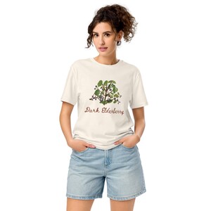以下が含まれることがあります： クリーム色のTシャツ。エルダーベリーの植物画が描かれており、緑の葉と濃い色の実が特徴です。「Dark Elderberry」の文字が筆記体でプリントされています。モデルは青いデニムのショートパンツを着用しています。