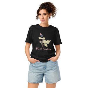 Op de afbeelding: Zwart T-shirt met een botanische illustratie van een braamplant en de tekst "Black Raspberry". Het model draagt een spijkerbroekshort. Het T-shirt is gemaakt van een zacht materiaal.