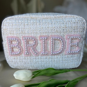 May include: White tweed cosmetic bag with the word "BRIDE" spelled out in pink letters with pearl accents.