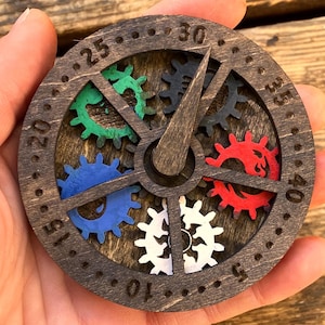 May include: A circular dark brown wooden game tracker featuring a rotating pointer and five colored gears in green, black, red, white, and blue. The outer rim is engraved with numbers from 5 to 40 in increments of five, arranged in a clock-like layout.