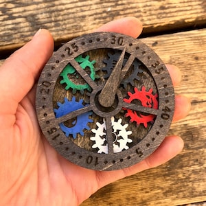 May include: A wooden clock with a dark brown finish. The clock face features a series of colorful gears in green, blue, red, and white. The clock hands are made of wood. The clock is held in a hand.