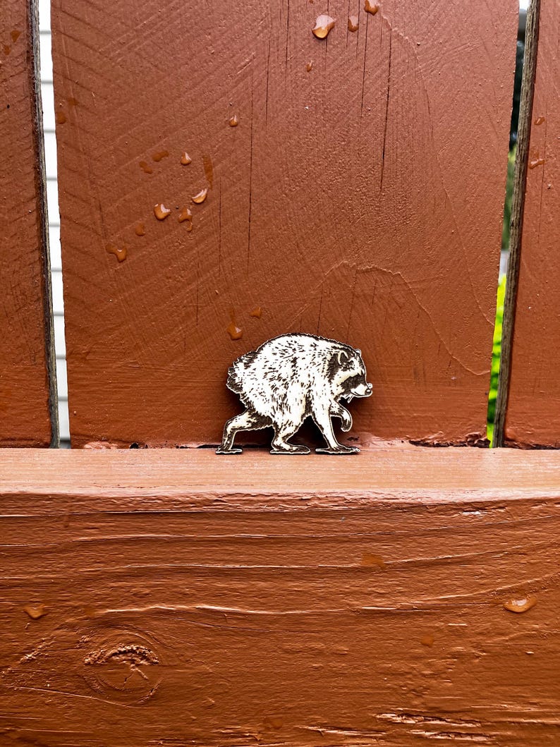 May include: A small, flat, wooden cutout shaped like a raccoon in a walking pose. The raccoon figure features detailed, dark-etched lines depicting fur texture, facial features, and a striped tail, creating a high-contrast black and light wood design.