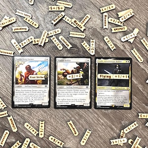 Peut inclure: Trois cartes Magic: The Gathering avec des personnages et des capacités, entourées de jetons en bois avec des effets de jeu comme "Vol" et "Première Frappe". Les cartes sont sur une surface en bois.