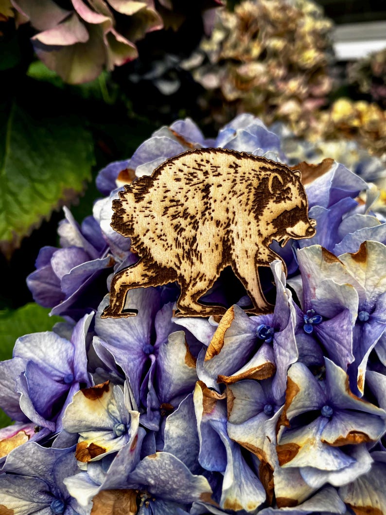 May include: A light-colored wooden cutout shaped like a raccoon, featuring dark, burnt-in details that define its fur texture, facial mask, and posture. The raccoon is depicted in a walking stance, showcasing intricate wood-burned lines throughout its body.