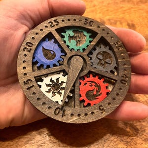 May include: A circular wooden game piece with a rotating dial and coloured gear designs. The gears are blue, green, grey, red, and white. The dial has numbers from 0 to 45, and a central pointer. The piece is held in a hand.