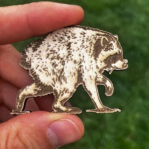 May include: A laser-cut wooden cutout shaped like a raccoon in a walking pose. The light-colored wood features dark, burnt-in details that define the raccoon's fur texture, facial features, and characteristic eye mask, creating a high-contrast design.
