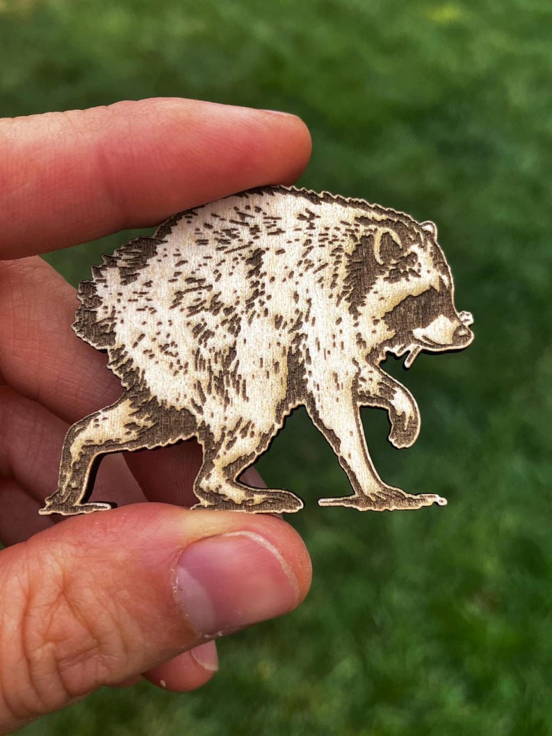 May include: A laser-cut wooden cutout shaped like a raccoon in a walking pose. The light-colored wood features dark, burnt-in details that define the raccoon's fur texture, facial features, and characteristic eye mask, creating a high-contrast design.