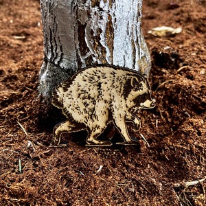 May include: A flat, laser-cut wooden cutout of a raccoon in a walking pose. The wood features dark, burned-in details that define the raccoon's fur texture, facial markings, and body shape, creating a high-contrast, detailed illustration on the light wood.