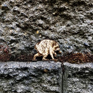May include: A flat, laser-cut wooden cutout of a raccoon in a walking pose, featuring detailed etched fur textures and dark facial markings. The light-colored wood piece shows a side profile of the animal with its head turned toward the right.