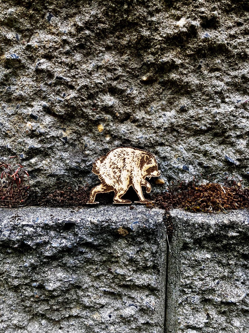 May include: A flat, laser-cut wooden cutout of a raccoon in a walking pose, featuring detailed etched fur textures and dark facial markings. The light-colored wood piece shows a side profile of the animal with its head turned toward the right.