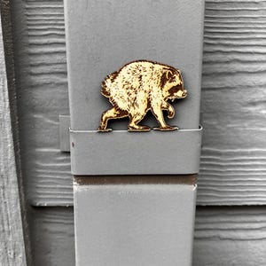 May include: A light brown wooden cutout of a raccoon in a walking pose, featuring detailed laser-engraved fur texture and facial features. The raccoon figure is positioned horizontally against a flat, grey, vertical metal surface with a smooth finish.