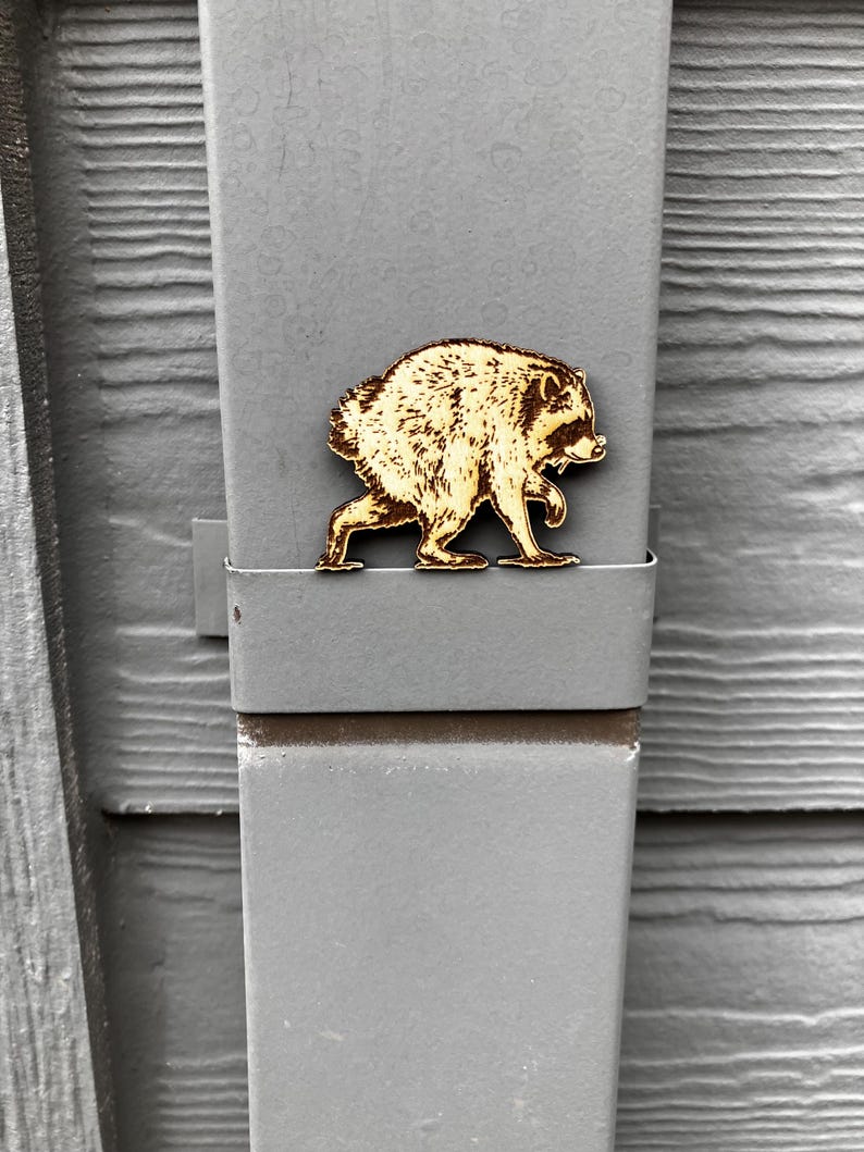 May include: A light brown wooden cutout of a raccoon in a walking pose, featuring detailed laser-engraved fur texture and facial features. The raccoon figure is positioned horizontally against a flat, grey, vertical metal surface with a smooth finish.