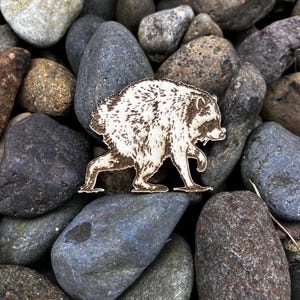 May include: A laser-cut wooden raccoon figurine featuring detailed dark brown etched fur patterns and facial markings. The raccoon is depicted in a side profile walking stance with its head turned slightly toward the viewer, showcasing its distinct mask.
