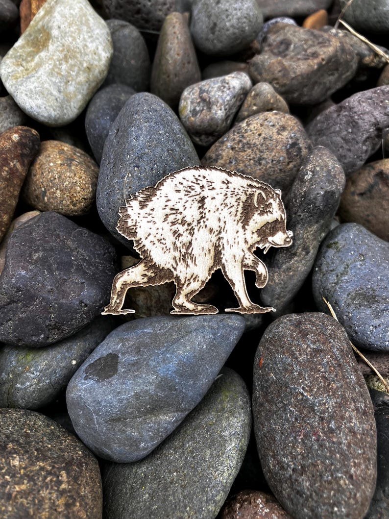 May include: A laser-cut wooden raccoon figurine featuring detailed dark brown etched fur patterns and facial markings. The raccoon is depicted in a side profile walking stance with its head turned slightly toward the viewer, showcasing its distinct mask.