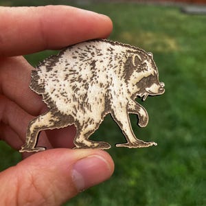 May include: A light-colored wooden cutout shaped like a raccoon in a walking pose. The design features dark, laser-engraved details highlighting the raccoon's fur texture, facial mask, and pointed ears, creating a high-contrast silhouette on the wood.