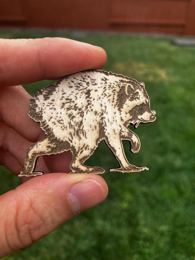 May include: A light-colored wooden cutout shaped like a raccoon in a walking pose. The design features dark, laser-engraved details highlighting the raccoon's fur texture, facial mask, and pointed ears, creating a high-contrast silhouette on the wood.