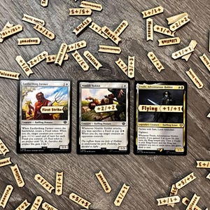 Pode incluir: Três cartas de Magic: The Gathering com fichas de madeira. As cartas apresentam personagens e habilidades como "First Strike" e "Flying". Fichas de madeira com efeitos de jogo como "+1/+1" e "Lifelink" estão espalhadas.