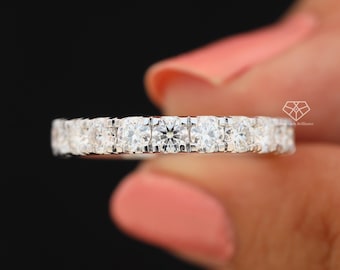 May include: A silver eternity ring with multiple round-cut, clear gemstones. The ring is held against a black background, showcasing the sparkle of the stones. The ring is a classic design, suitable for various occasions.