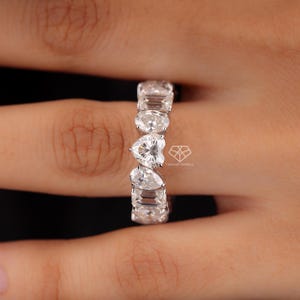 May include: A white gold ring with a series of clear, sparkling gemstones. The gemstones are cut into various shapes, including oval, heart, pear, and emerald cuts. The ring is set against a neutral background.