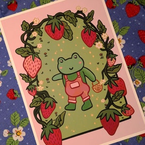 May include: A green frog wearing overalls stands in a field of strawberries. The frog is surrounded by strawberry plants with red strawberries and white flowers. The background is a light green color with white sparkles.