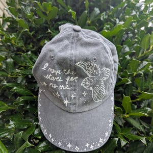 May include: A grey baseball cap featuring white hand-painted details including a flying owl, scattered stars, and the text "I move the stars for no one" written in cursive script across the front panel, with a star border along the curved brim.