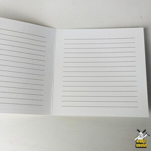 May include: Open notebook with lined pages. The notebook is white with black lines and is open to reveal two facing pages. The notebook is on a white surface. The lower right corner has a logo with a rabbit and the words "Rabbit Banana".