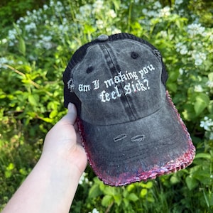 May include: Distressed grey baseball cap with a black mesh back and a curved peak. The cap features the embroidered text "am I making you feel sick?" in white. The peak is accented with a dark red sequined trim.