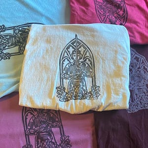 May include: Four t-shirts featuring a black and white print of a figure in a stained glass window. The figure is holding a sword and is surrounded by flowers. The t-shirts are in different colours: light blue, white, pink, and maroon.