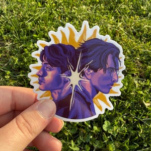 May include: A sticker featuring two side profiles of individuals with dark hair, facing away from each other. The portraits are in shades of purple and blue, set against a yellow sunburst design. The sticker is held against a backdrop of green grass.