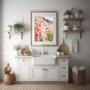 May include: A watercolor painting of a colorful Italian village with pink buildings and green trees, framed in a light wood frame and hanging on a white wall above a white kitchen sink.