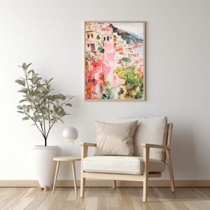 May include: A watercolor painting of a colorful Italian village with pink buildings, green trees, and a blue ocean in the background. The painting is framed in a light wood frame and is hanging on a white wall.