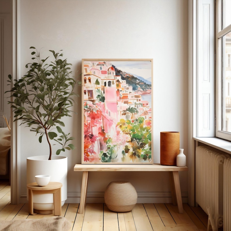 May include: A watercolor painting of a colorful Italian village on a hillside overlooking the sea. The painting is framed in a light wood frame and is displayed on a wooden bench with a woven basket and a vase.